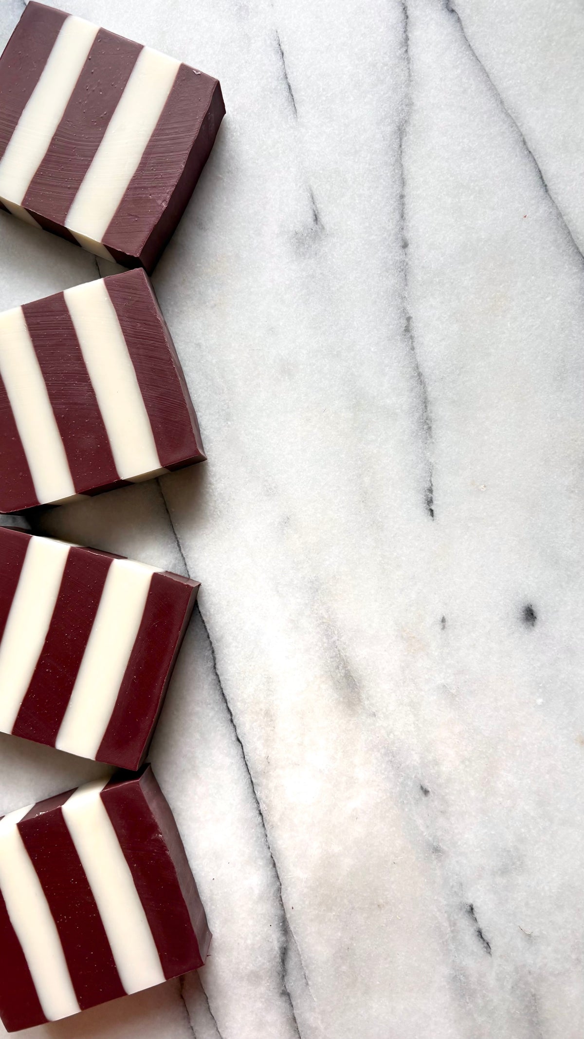 Four rectangular bars of striped soap in alternating deep burgundy and off-white, arranged diagonally on a white marble surface with gray veining.