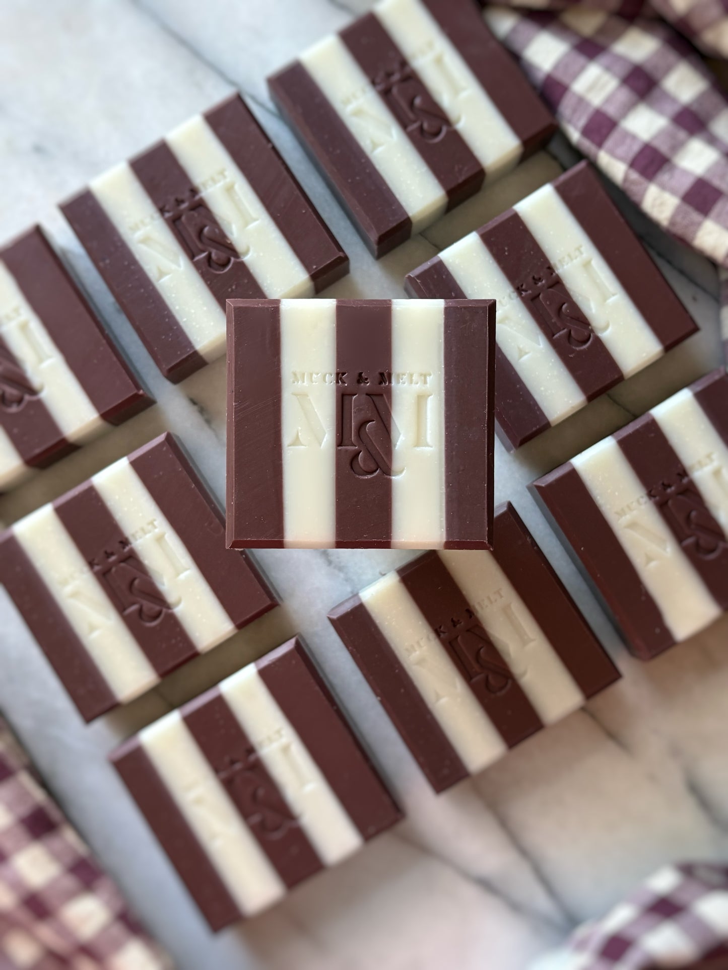 A top-down view of burgundy plum and white striped soaps  arranged on a light marble surface with a purple-and-white gingham cloth in the corners. The center bar is embossed with the brand name 'Muck and Melt' and a monogram.