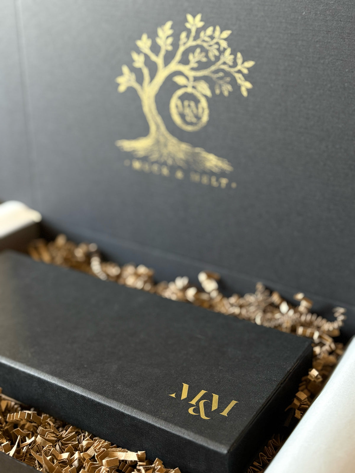 A close-up of a black presentation box with a gold tree logo and the initials 'M&M' on the lid, resting in shredded brown paper packing material inside an open matching box.