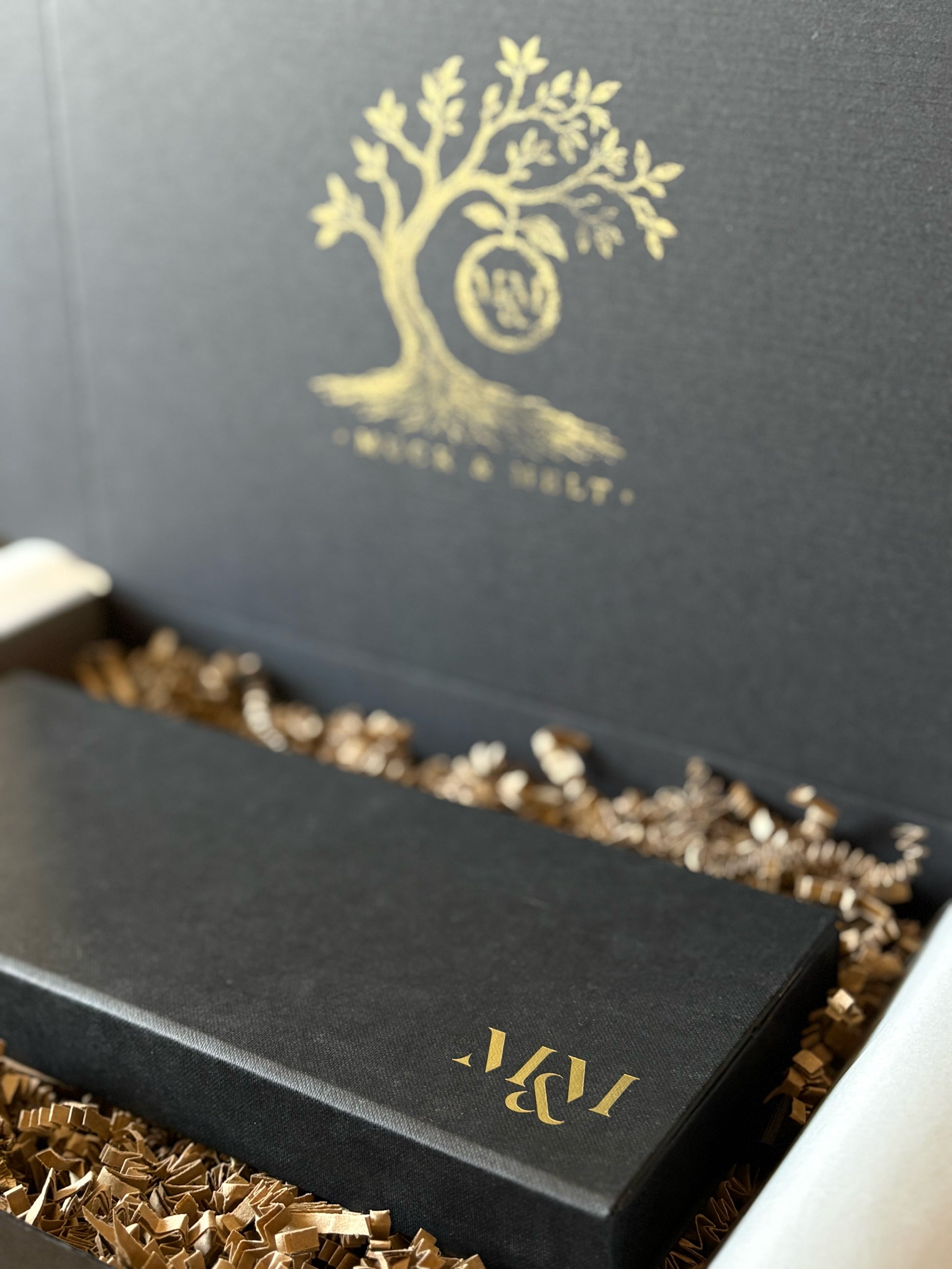 A close-up of a black presentation box with a gold tree logo and the initials 'M&M' on the lid, resting in shredded brown paper packing material inside an open matching box.