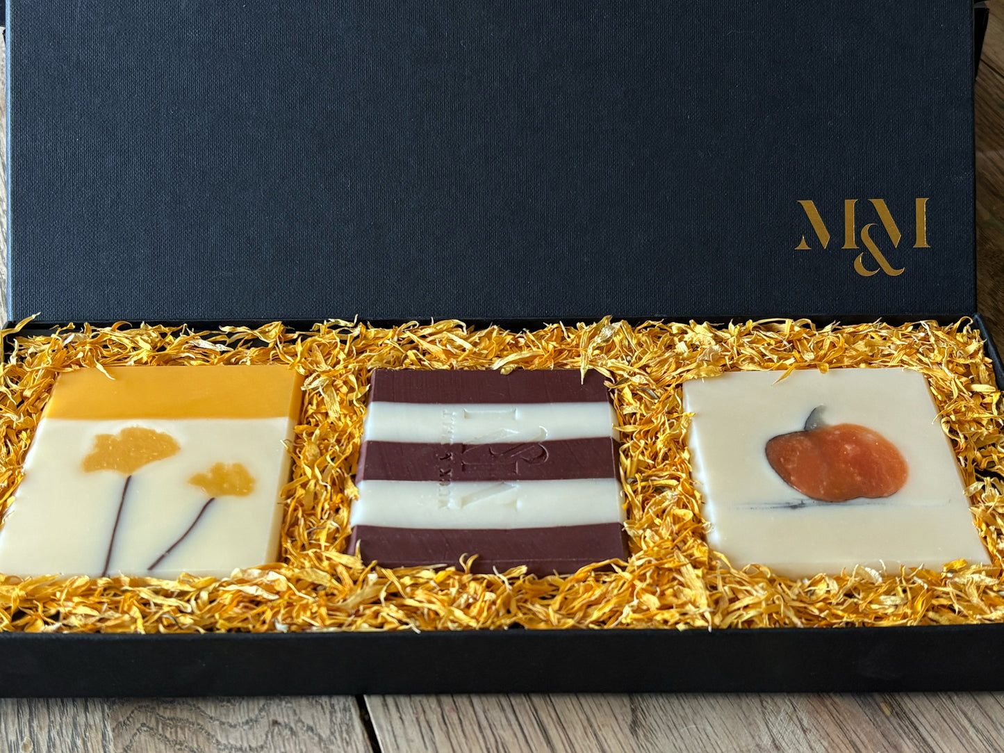 Three artisanal square soaps displayed in a black gift box lined with golden calendula petals. The left soap is white with yellow ginkgo leaves, the center soap has dark burgundy and white horizontal stripes and the right soap is white with a small orange pumpkin.