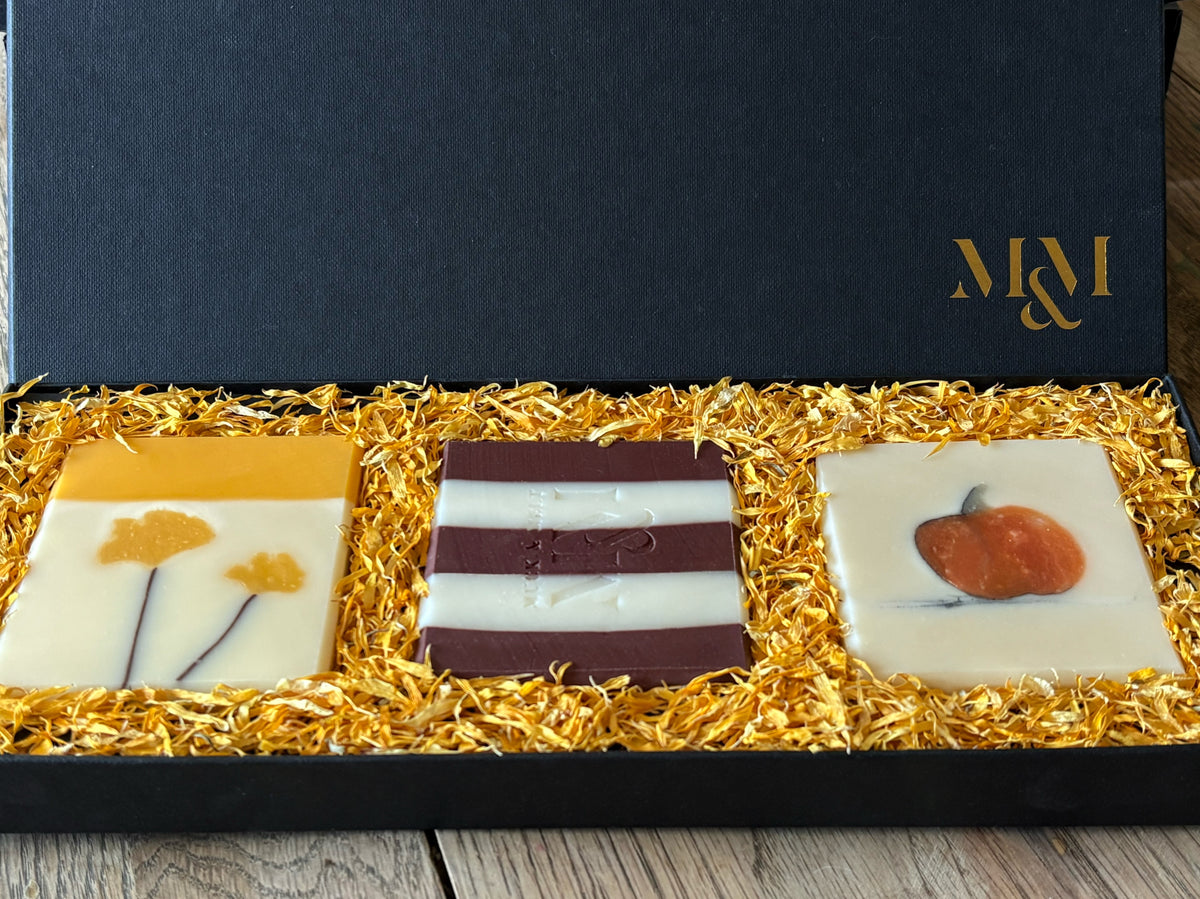 Three artisanal square soaps displayed in a black gift box lined with golden calendula petals. The left soap is white with yellow ginkgo leaves, the center soap has dark burgundy and white horizontal stripes and the right soap is white with a small orange pumpkin.