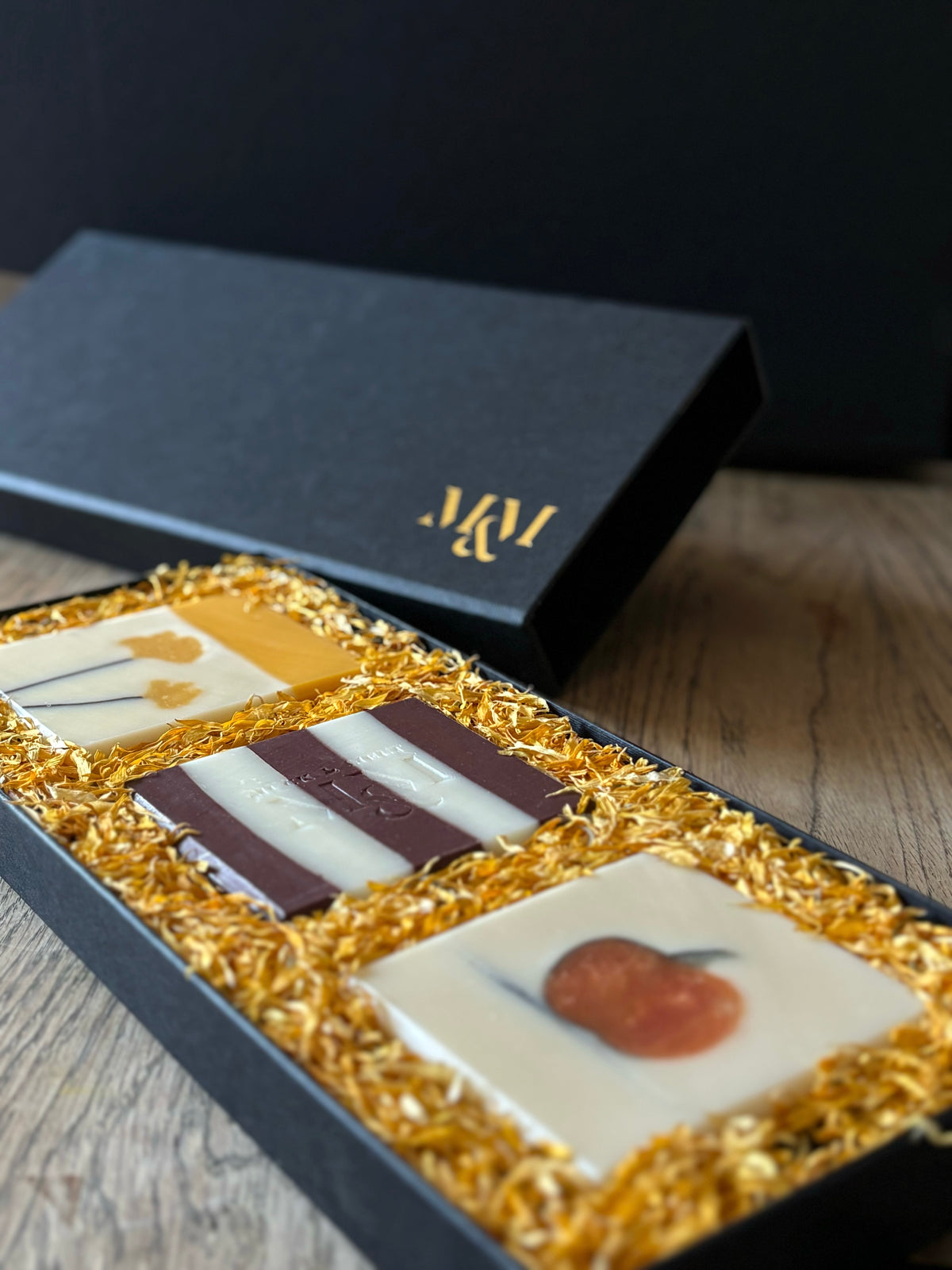 A luxury black gift box containing 3 handmade natural soaps. a cream soap with a yellow top and a Ginkgobiloba leaf image. A plum burgundy and white striped soap. a cream soap withthe image of a pumpkin in the centre in the style of a sketch. nestled in yellow calendula petals.