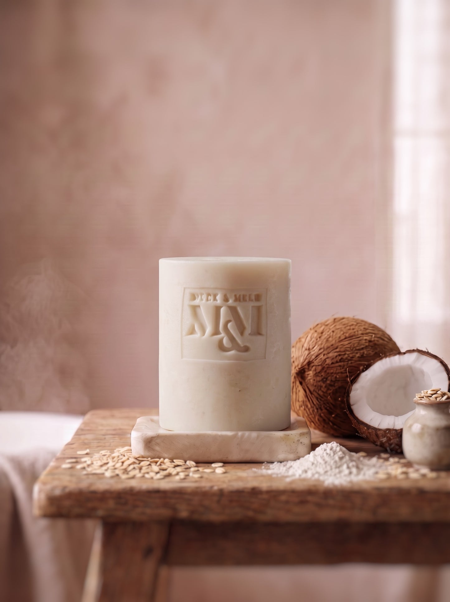 A white soap with a monogram logo embossed on the front, displayed on a rustic wooden table alongside a halved coconut, oat flakes, and a small jar against a soft, warm beige background.