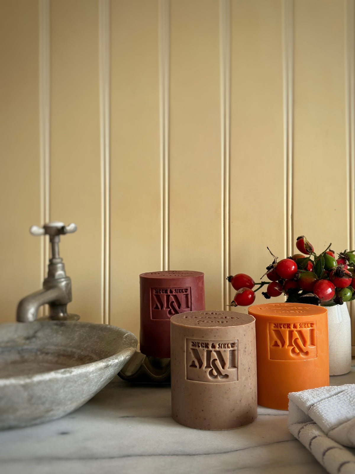 Three large cylindrical soaps embossed 'MUCK & MELT' branding, displayed on a marble countertop next to a rustic sink, a folded towel, and red rosehips against a beige panelled wall.