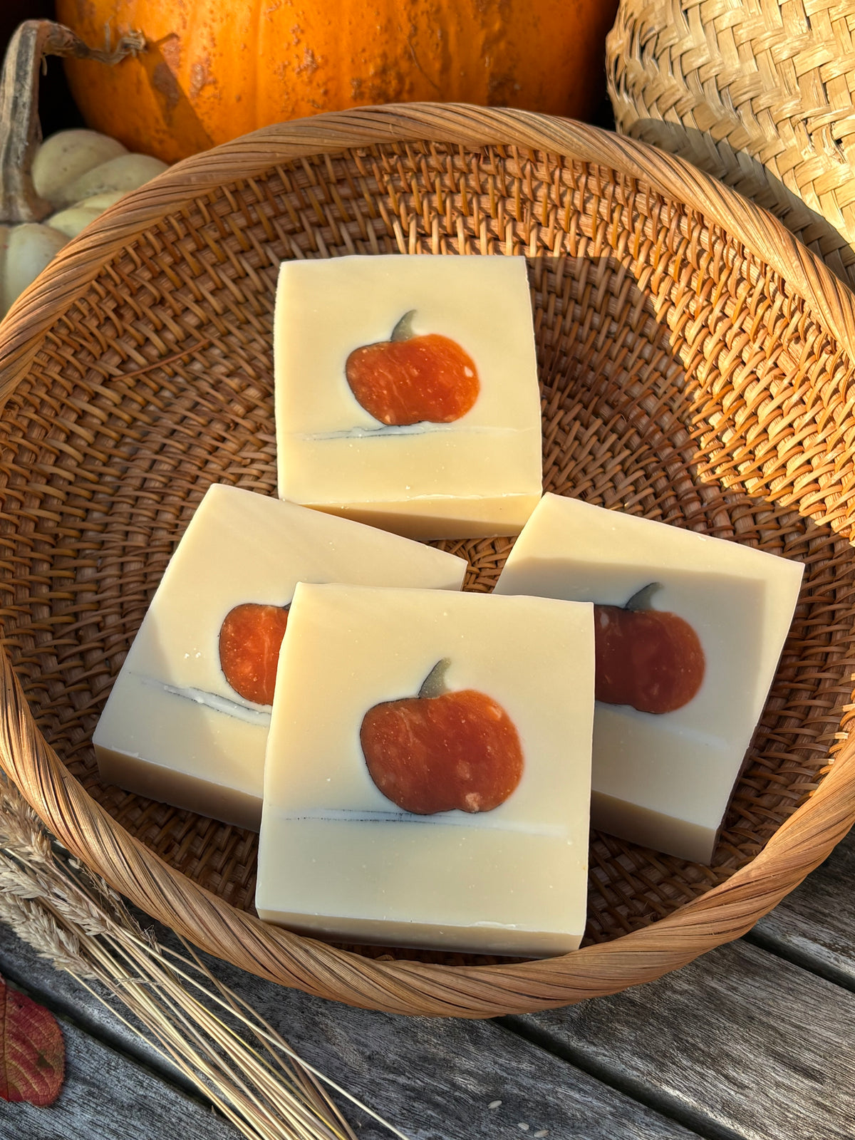 Four rectangular bars of pale cream-colored soap with a printed orange pumpkin design on top, arranged inside a round woven wicker basket. Pumpkins and a rustic wooden surface are visible in the background.