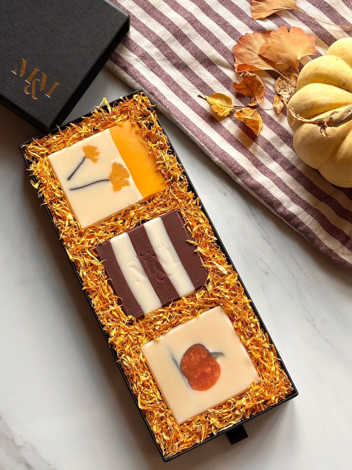 A black gift box filled with three artisanal handmade soaps nestled in calendula petals displayed on a white marble surface next to a striped cloth and dried autumn leaves with a pumpkin.