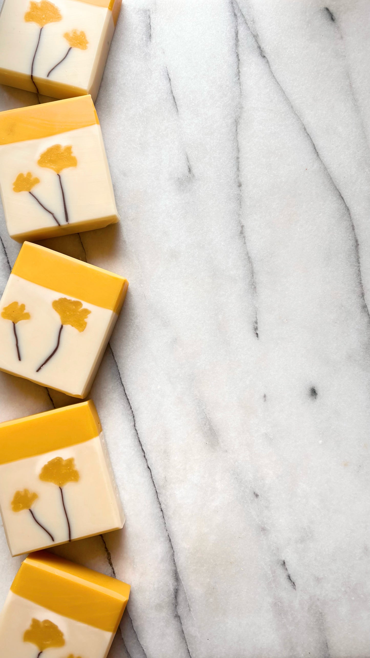 Five rectangular bars of soap arranged vertically along the left side of a white marble surface. Each soap has a warm yellow top half, a cream lower half, and a yellow flower with dark stems on the front.