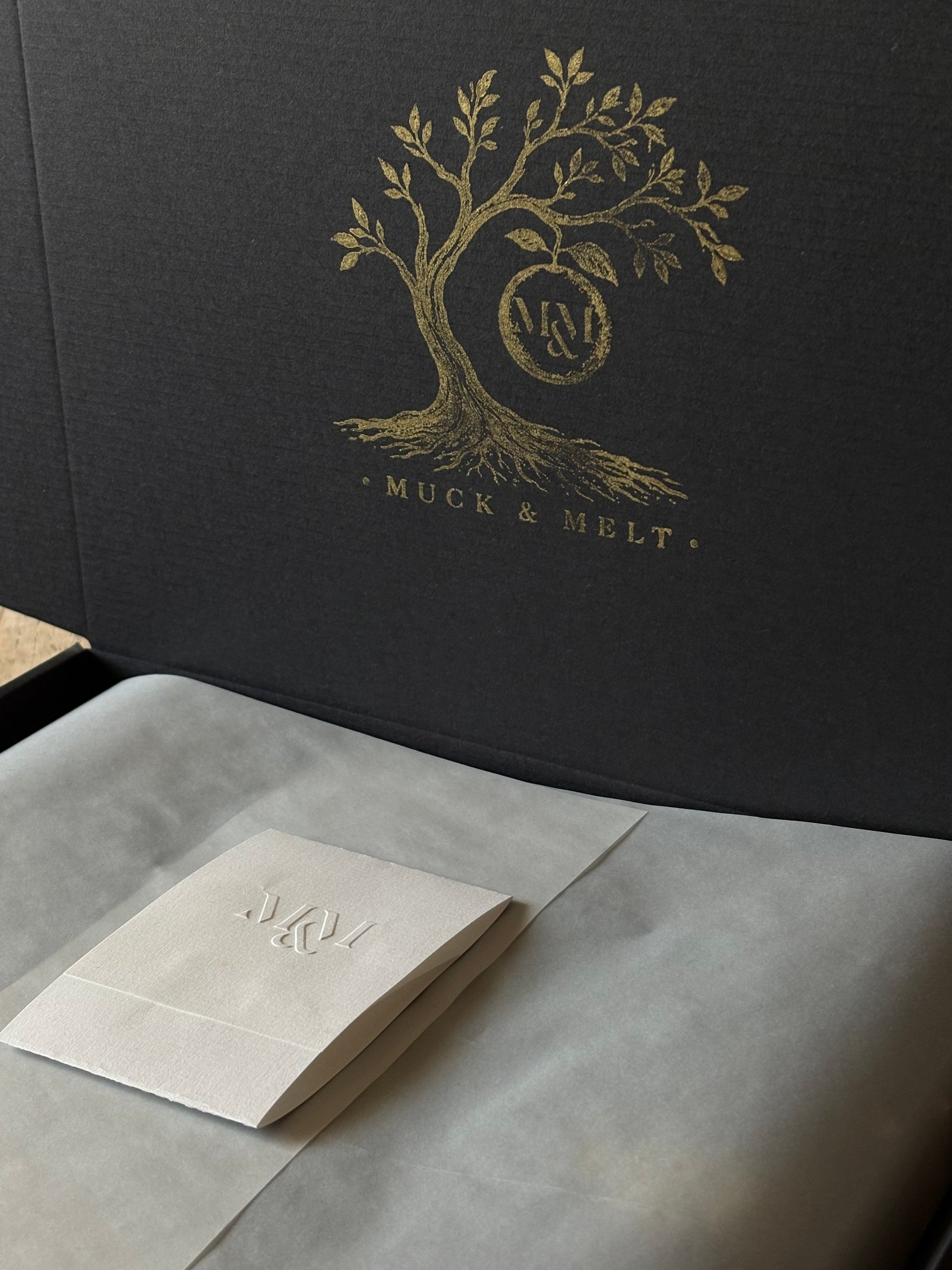 A close-up of a black textured presentation box with a gold foil tree logo and the words 'MUCK & MELT' on the inside lid, with a folded white tissue or paper insert resting on a light gray surface in front.