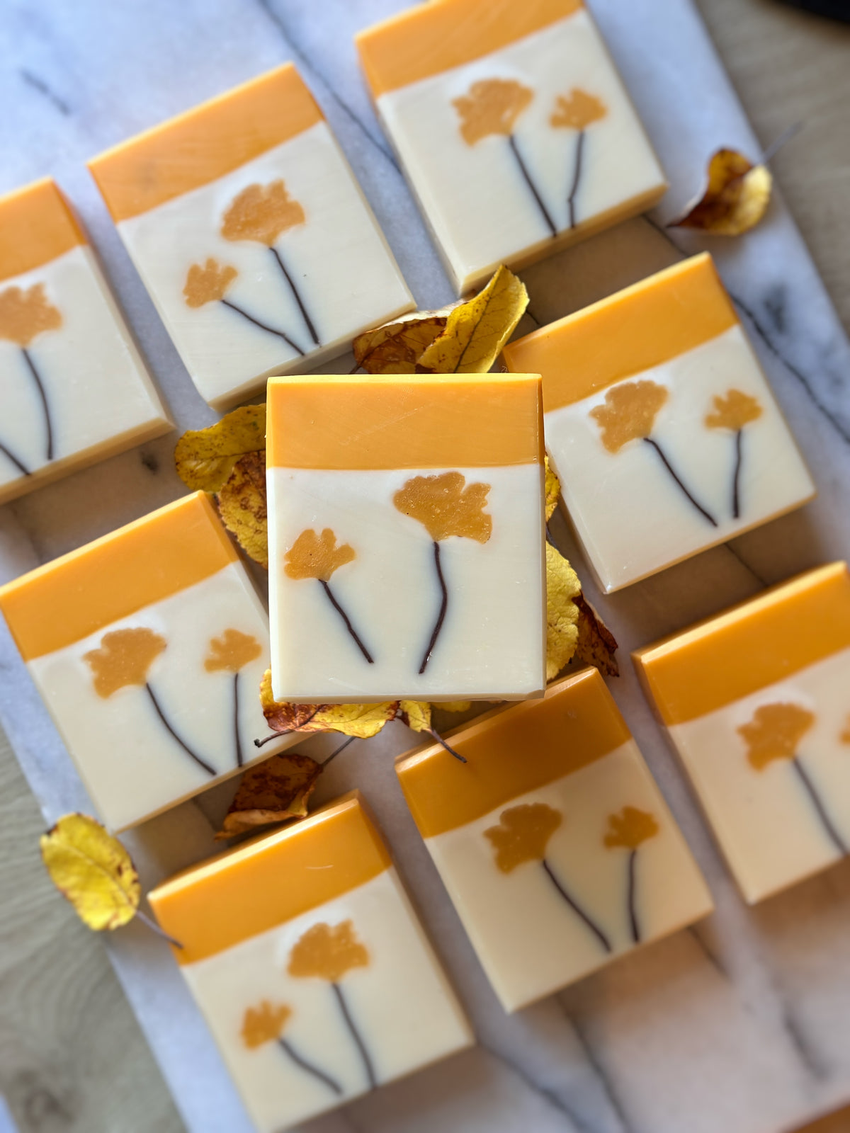 A top-down view of multiple rectangular bars of soap arranged on a light marble surface, with one central bar highlighted. The soap has a warm cream body, a bright yellow top band, and printed mustard-yellow and dark brown Ginkgo leaf motifs.