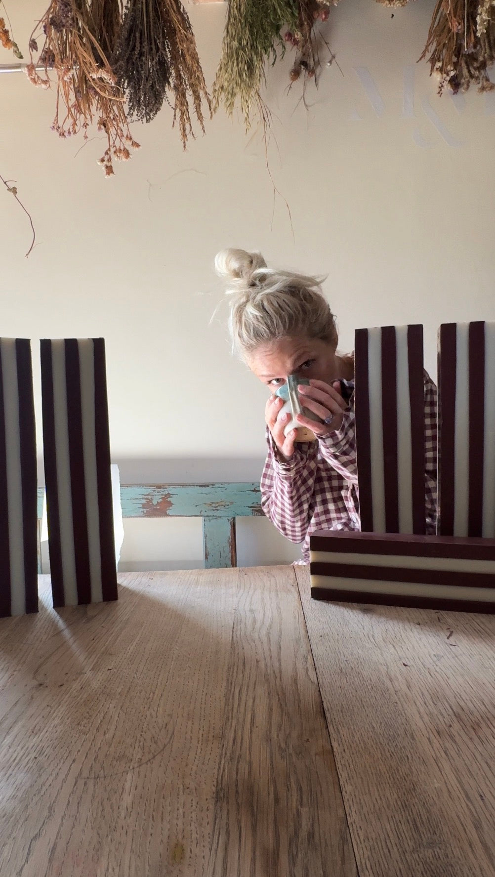 Lizzi in her pyjamas with blonde hair in a bun peeks over blocks of burgundy/ plum striped soap blocks holding a cup of coffee in a rustic room with a wooden floor and dried flowers hanging from the ceiling.